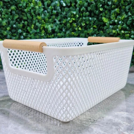 Cesto organizador de plástico branco com alças de bambu vazado visto de frente, fundo de folhagem