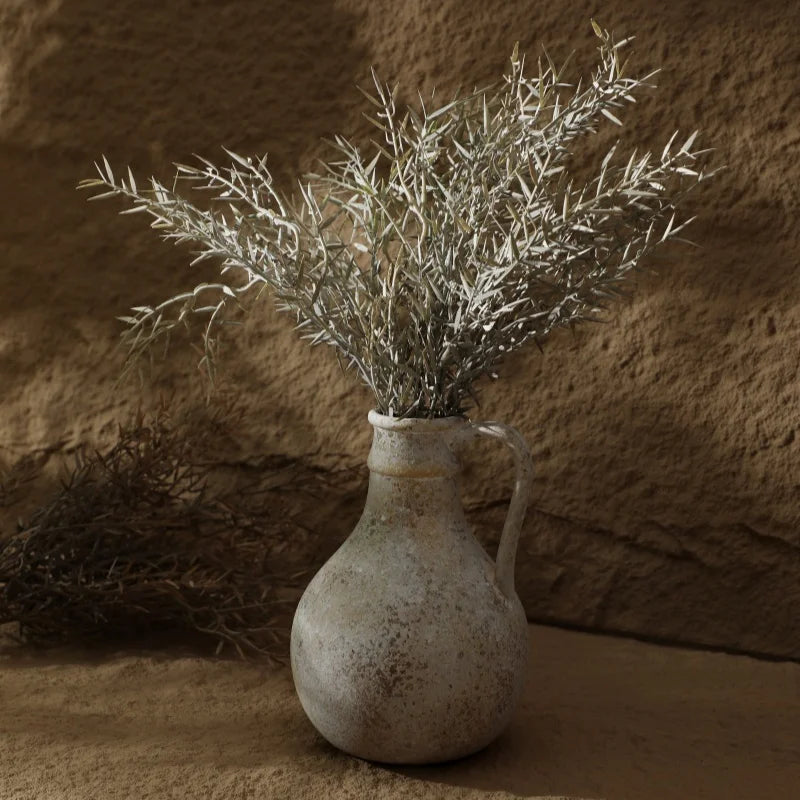 Vaso Decorarti em cerâmica rústica com galhos secos sobre superfície de areia e parede de pedra ao fundo