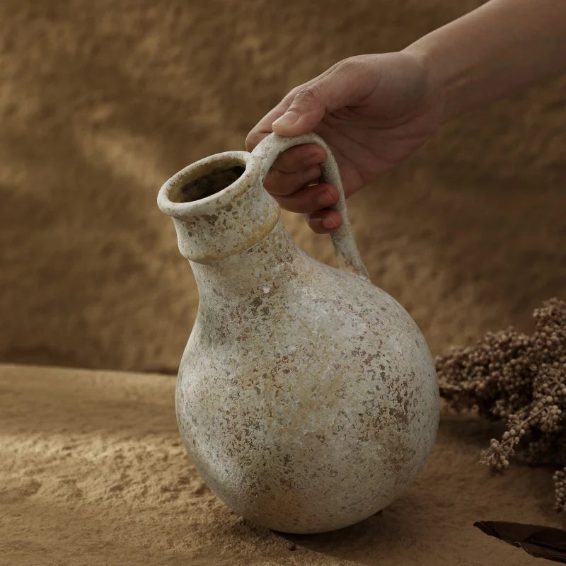 Mão segurando o Vaso Decorarti pela alça lateral, destacando a textura manchada cinza-bege da cerâmica